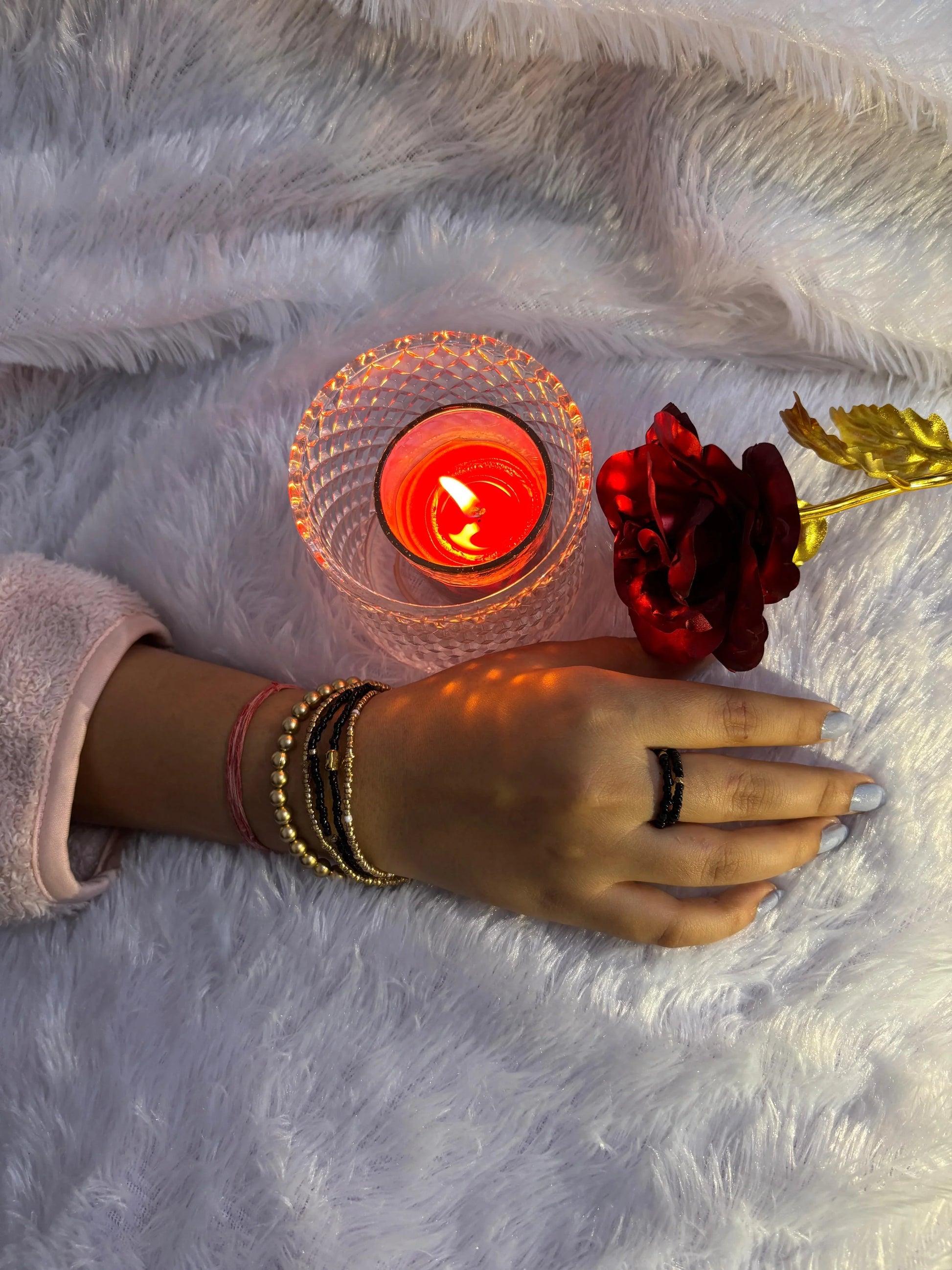 Hand with bracelets holding a red rose next to a lit candle on a fluffy white surface