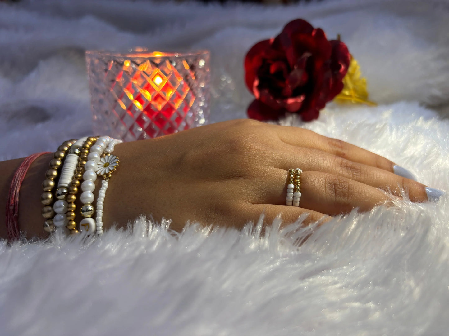 Hand wearing multiple bracelets and a ring on a fluffy white surface with a candle and flowers in the background.